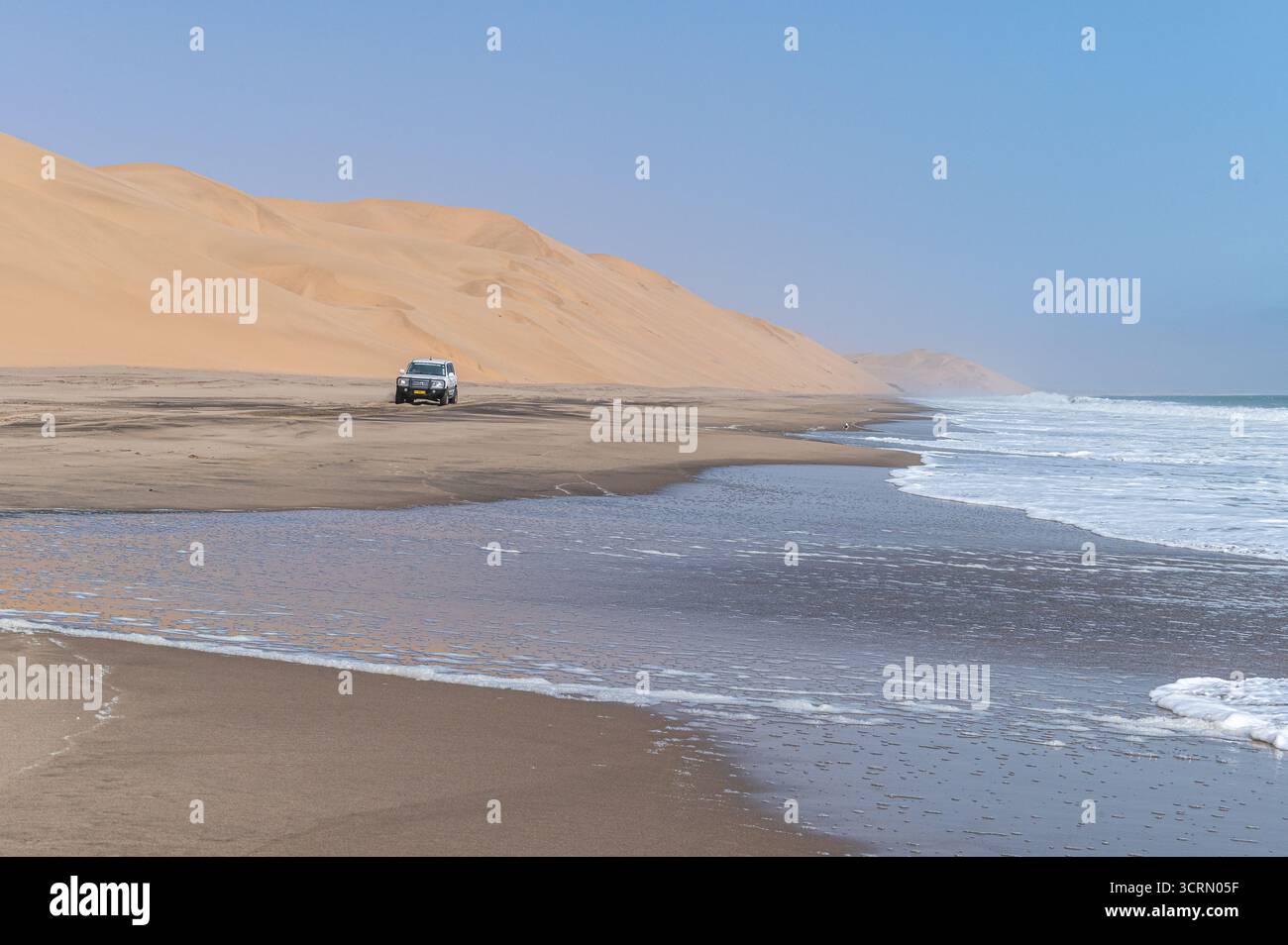 Lone 4x4 SUV, der von den Namib-Wüsten-Dünen an einem schmalen Küstenstrand in den Schatten gestellt wird, Atlantik-Surf auf der rechten Seite, wo die riesige Wüste auf das Meer trifft, Sandwich Harbour, Namibia. Stockfoto