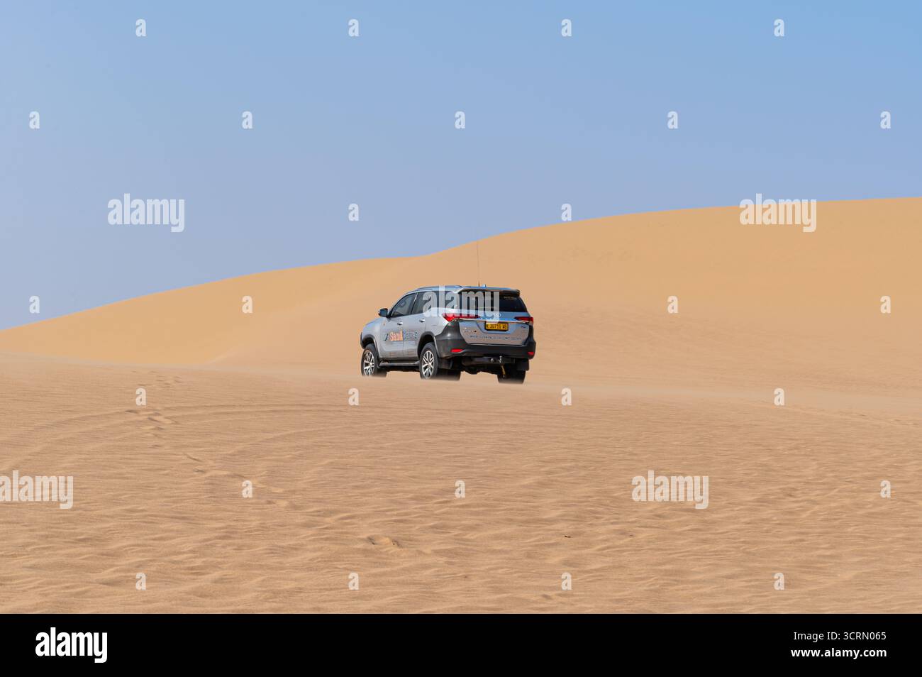 Rückansicht des weißen Toyota Fortuner SUV, der steile, goldene Namib-Düne, riesige Wüstenlandschaft und blauer Himmel im Hintergrund, Sandwich Harbour, Namibia. Stockfoto