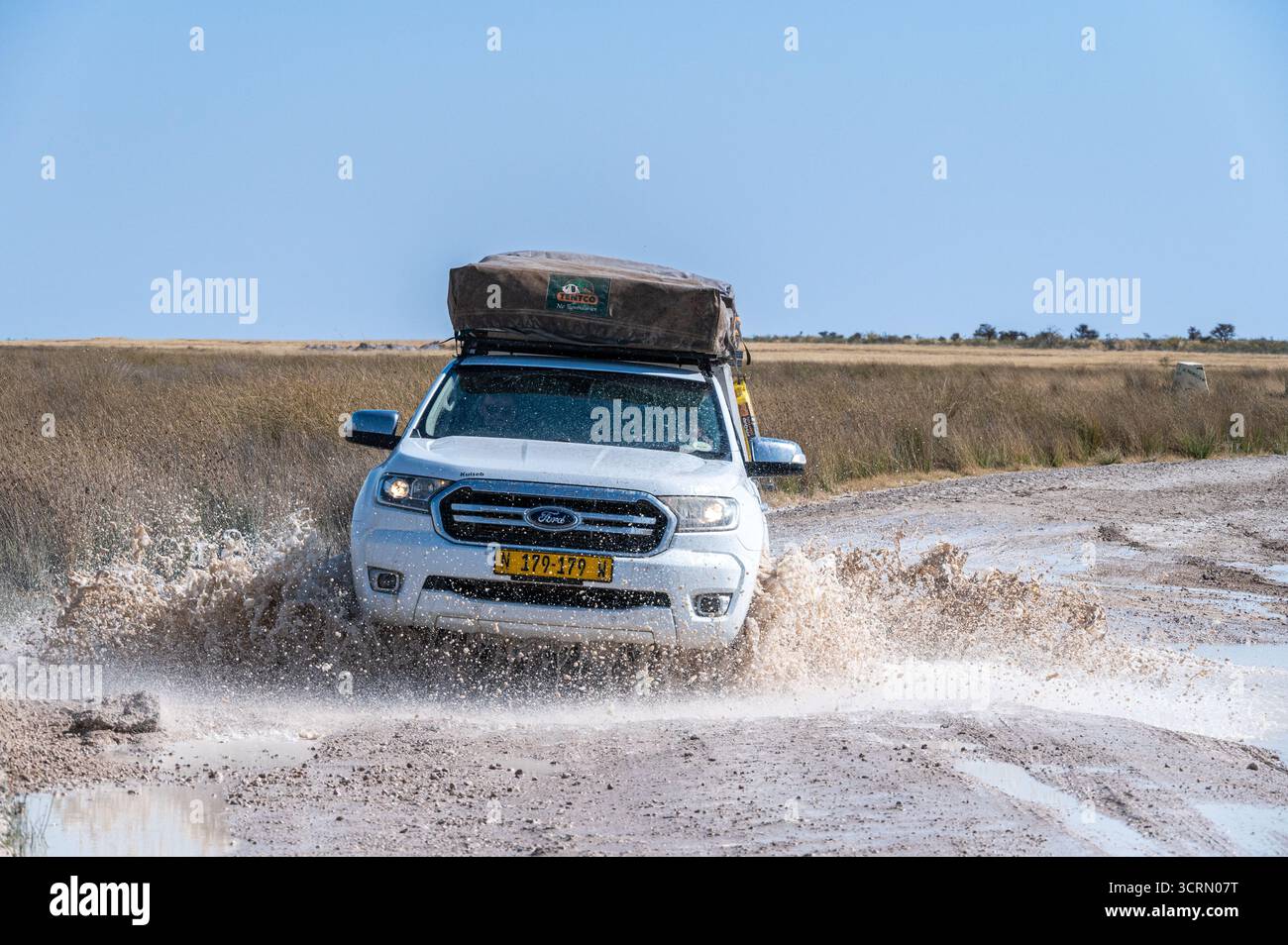 Frontale Held-Aufnahme eines weißen Doppelkabinen-Geländewagens mit zwei Dachzelten, die durch eine schlammige Salzpfanne platzen, im Etosha-Nationalpark, Namibia Stockfoto