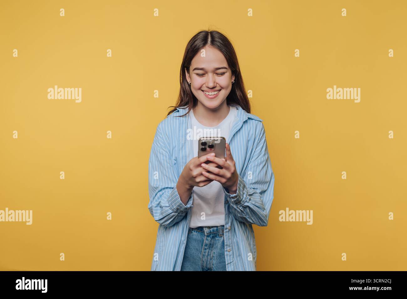 Eine lächelnde junge Frau mit einem gestreiften Hemd interagiert mit ihrem Smartphone vor einem hellgelben Hintergrund Stockfoto