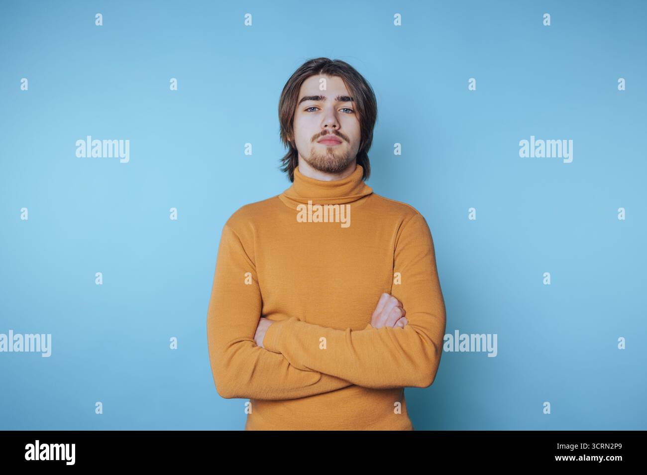 Ein selbstbewusster junger Mann in gelbem Rollkragenpullover steht vor blauem Hintergrund, während die Arme beim Fotoshooting in der Halle überkreuzt werden Stockfoto