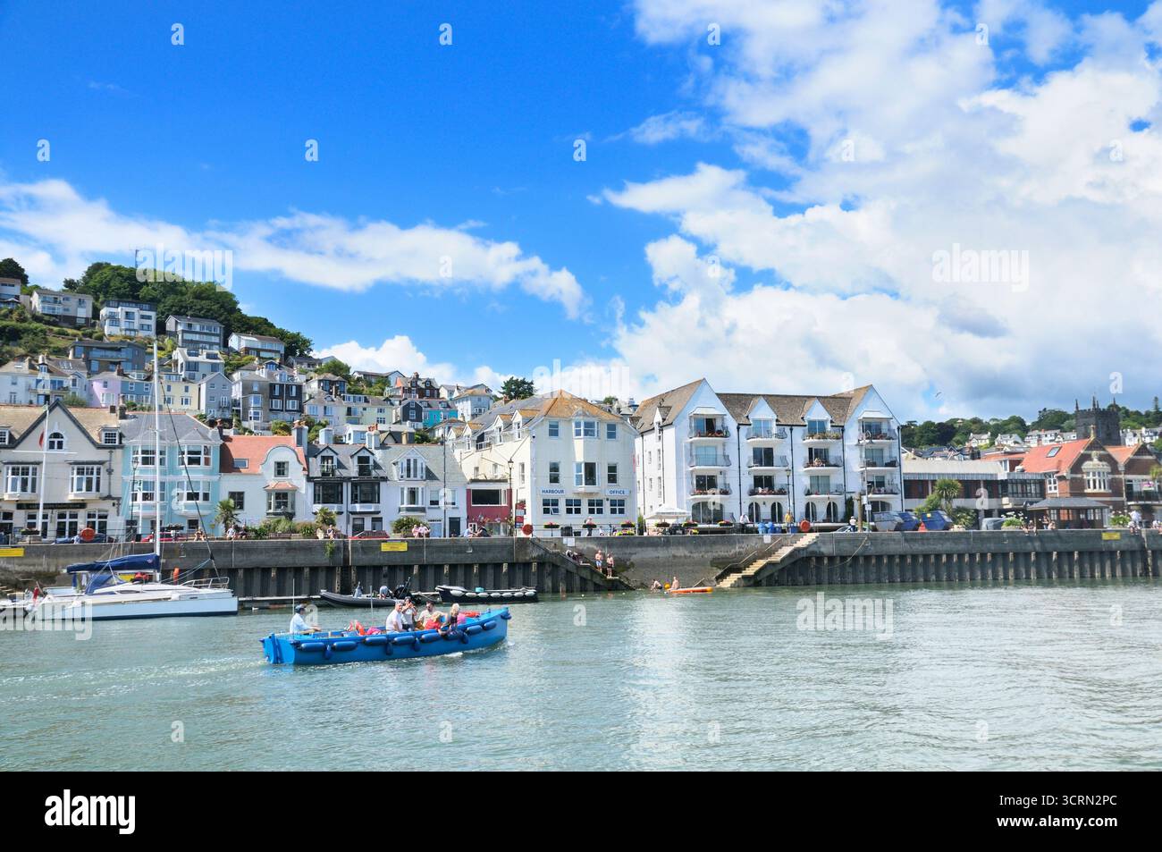 Ein Blick auf die Gebäude am Ufer entlang des Dartmouth's South Embankment im Sommer mit Touristen in einem Boot und am Kai, South Hams, Devon, England, Großbritannien Stockfoto