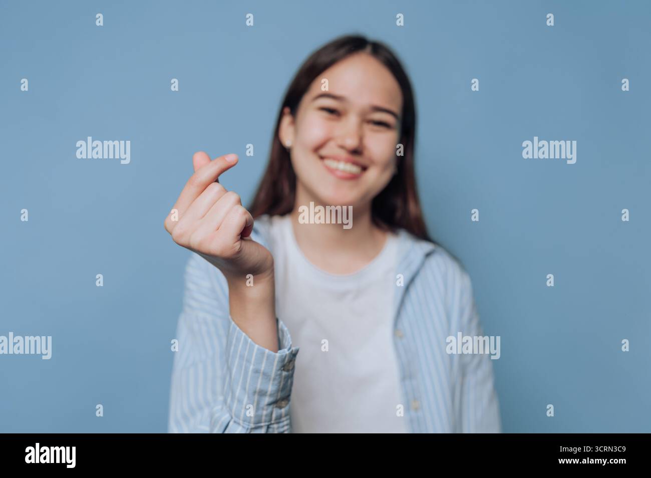 Eine fröhliche junge Frau zeigt mit ihren Fingern eine Herzgeste, die vor einem hellblauen Hintergrund steht. Ihr Lächeln strahlt Glück und Positivit aus Stockfoto