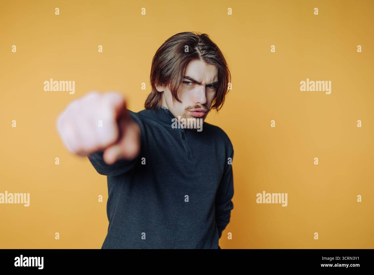 Ein junger Mann mit langen Haaren zeigt selbstbewusst mit dem Finger, der gegen eine hellgelbe Wand gerichtet ist. Er strahlt eine Mischung aus Entschlossenheit und Einstellung aus Stockfoto Ein junger Mann mit langen Haaren zeigt selbstbewusst mit dem Finger, der gegen eine hellgelbe Wand gerichtet ist. Er strahlt eine Mischung aus Entschlossenheit und Einstellung aus Stockfoto