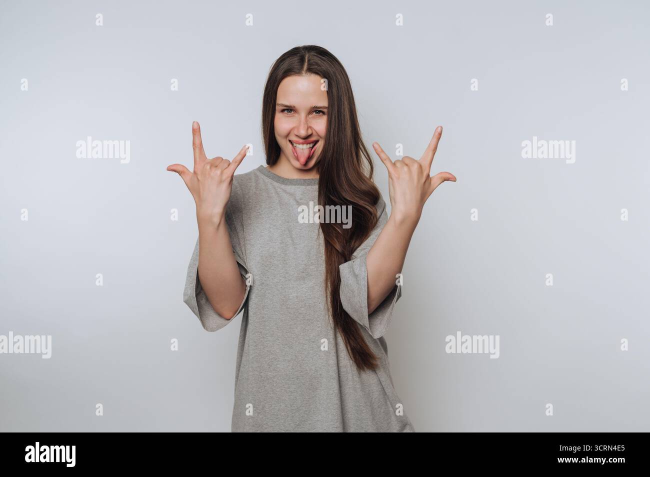 Eine junge Frau mit langen Haaren, die ein graues T-Shirt trägt, macht eine verspielte Rock-and-Roll-Handgeste, während sie ihre Zunge gegen einen schlichten Hintergrundier ausstreckt Stockfoto