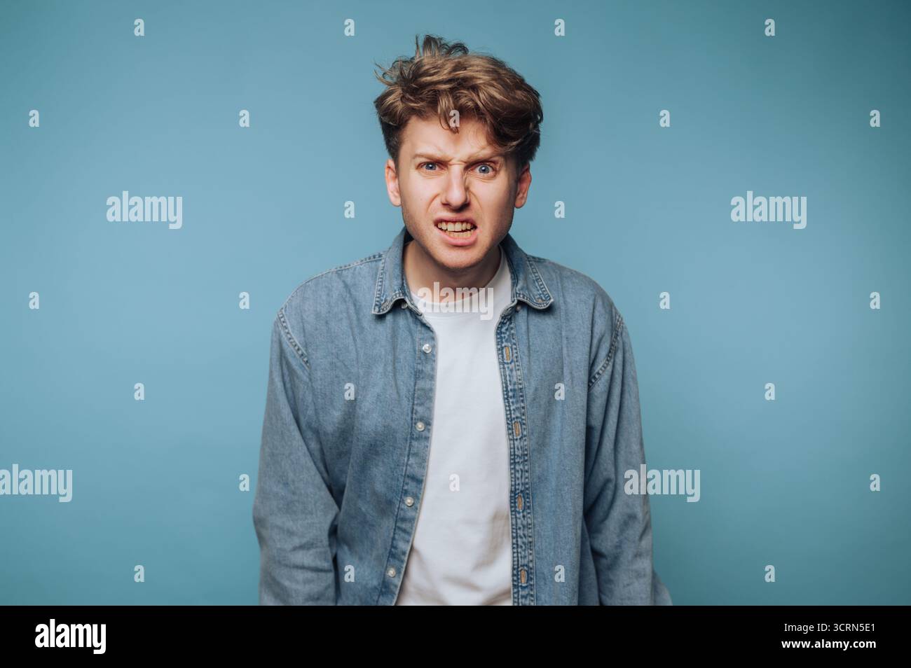 Junger Mann mit unordentlichen Haaren in Denim-Hemd mit wütendem Ausdruck vor blauem Hintergrund. Stockfoto