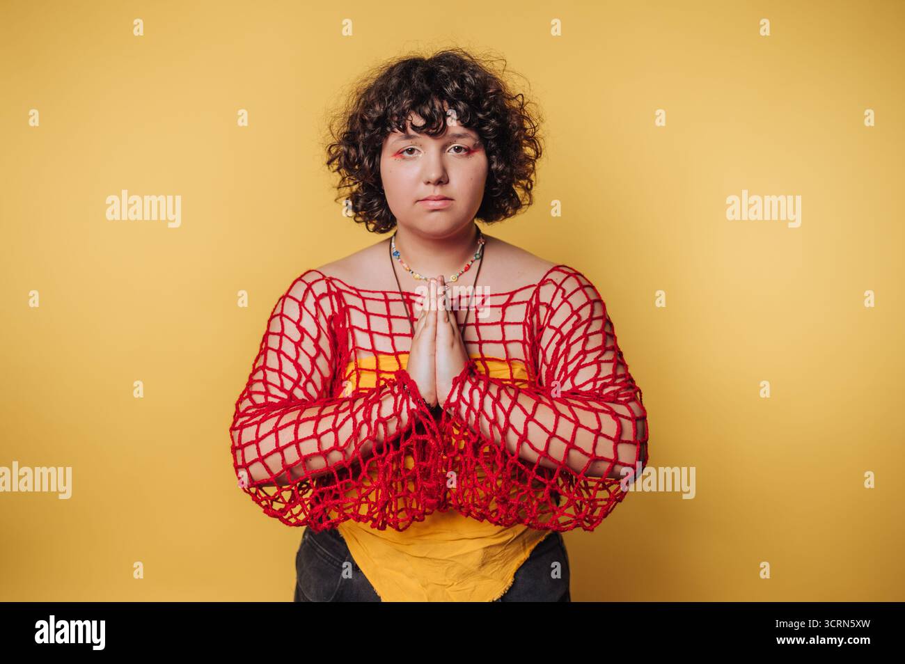 Eine Person mit lockigen Haaren in einem roten Netztop posiert mit Händen in einer Gebetsposition vor gelbem Hintergrund. Stockfoto