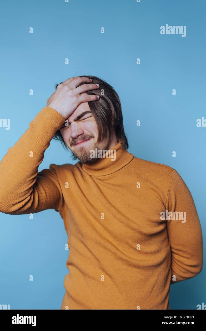Porträt eines jungen Mannes legt seine Hand auf seine Stirn und zeigt Gefühle von Stress oder Überforderung auf blauem Hintergrund Stockfoto