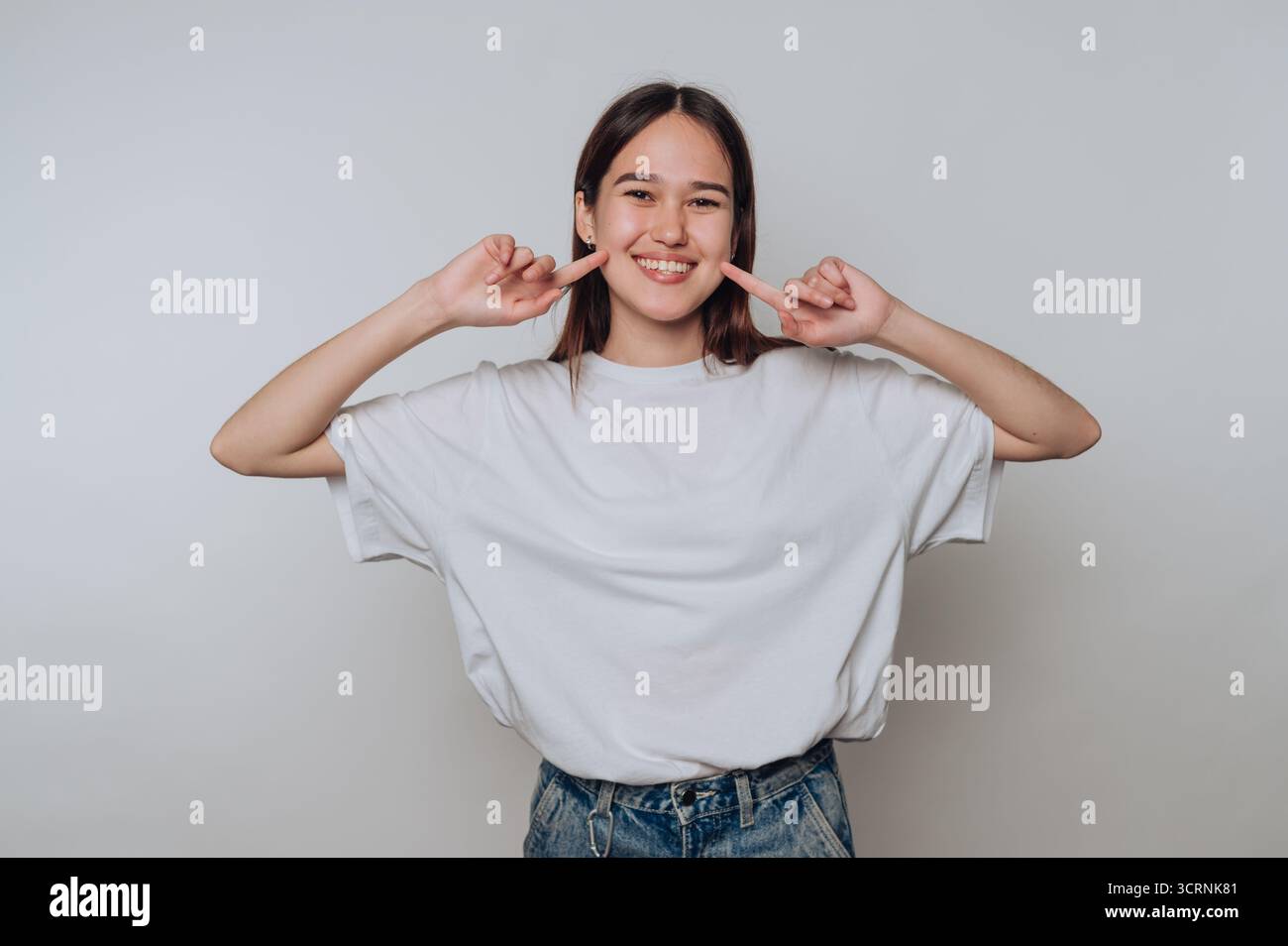 Eine junge Frau in lässiger Kleidung lächelt und zeigt auf ihre Wangen. Stockfoto