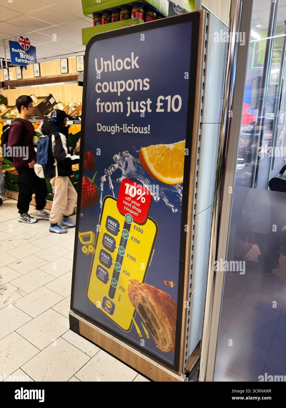 Manchester, Großbritannien - 25. September 2025: Die Käufer navigieren durch einen hellen Lidl-Lebensmittelgang, vorbei an einem kühnen Coupon-Schild und Gebäck-Bildern. Stockfoto