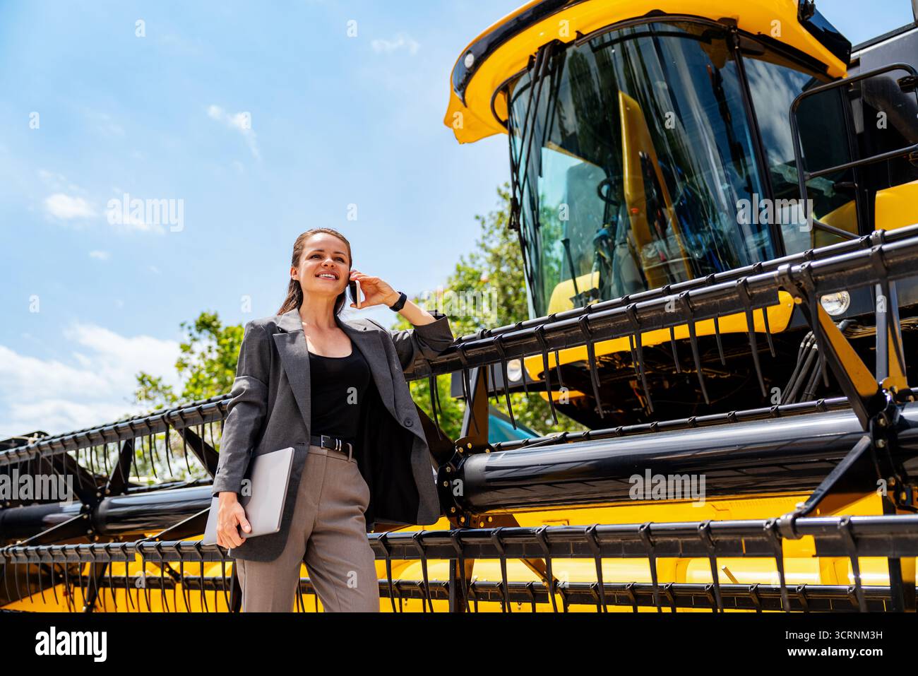 Selbstbewusste Frau in der Landwirtschaft, die sich auf den Mähdrescher stützt und am Telefon spricht Stockfoto