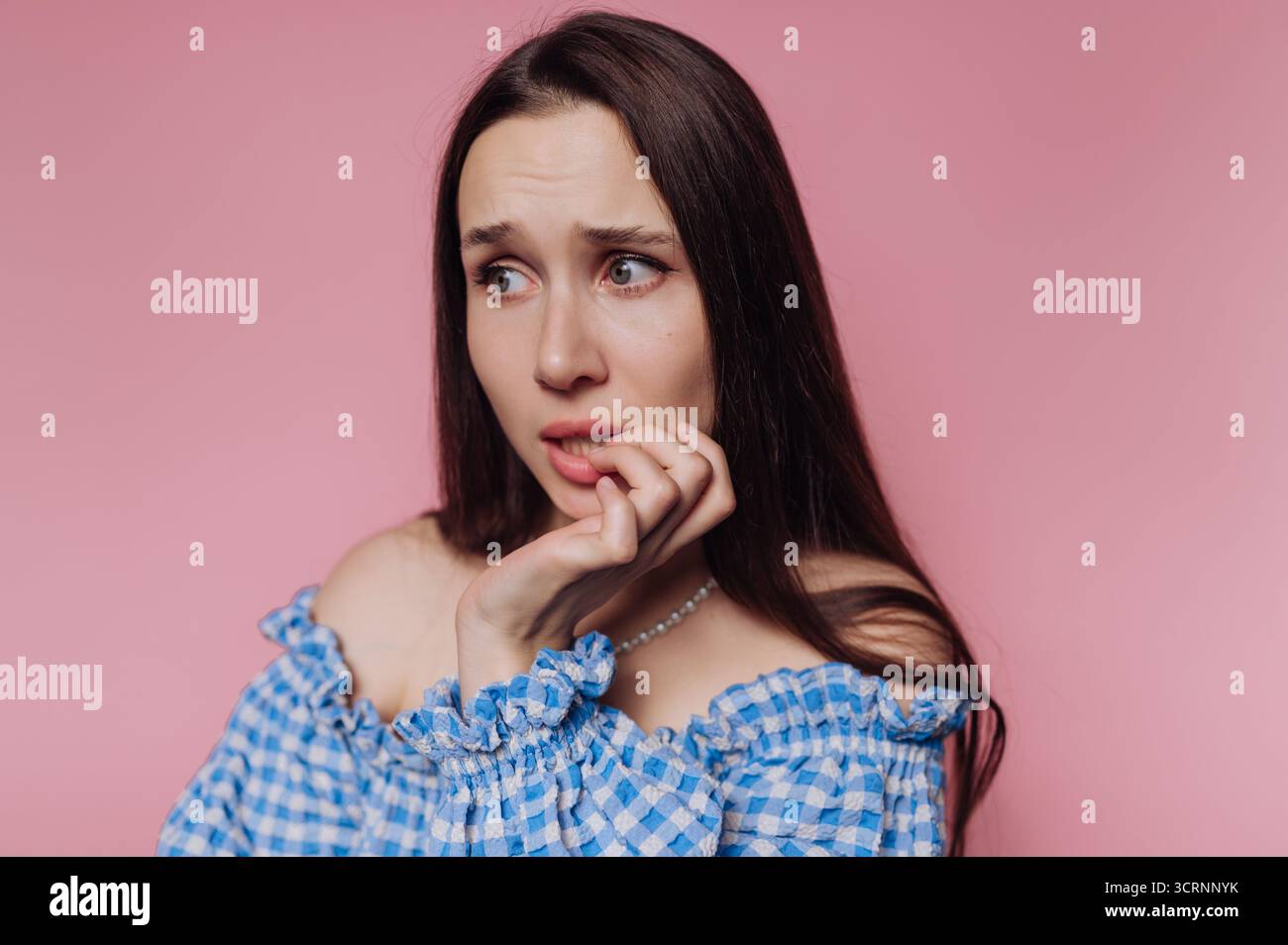 Eine besorgte Frau mit langen dunklen Haaren, die sich vor rosafarbenem Hintergrund in die Nägel beißt. Stockfoto