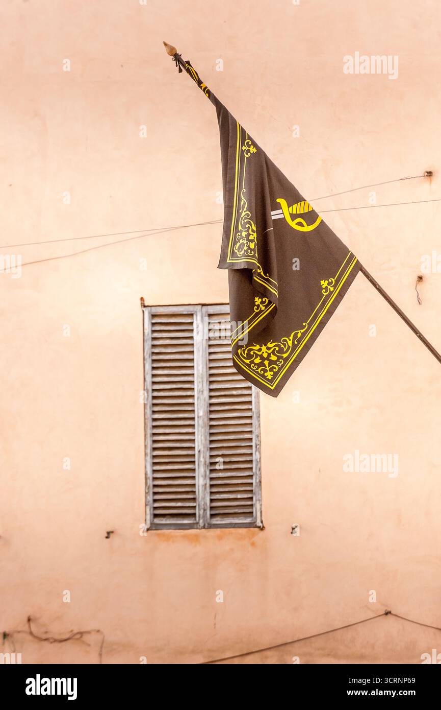 Braune Flagge im mittelalterlichen Stil mit gelben Details, die neben einem hölzernen Rolltor an einer pastellfarbenen Wand in Foligno, Umbrien, Italien winken Stockfoto