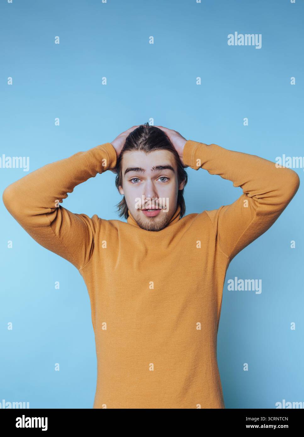 Porträt eines jungen Mannes mit einem Rollkragen blickt in die Ferne, Hände auf dem Kopf, reflektiert das Gewicht der Gedanken auf blauem Hintergrund Stockfoto