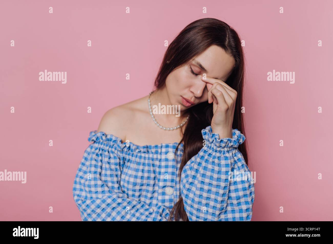 Eine junge Frau mit langen Haaren trägt ein blau-weiß kariertes Off-Shoulder-Kleid. Sie berührt ihre Stirn und scheint Unbehagen auszudrücken o Stockfoto