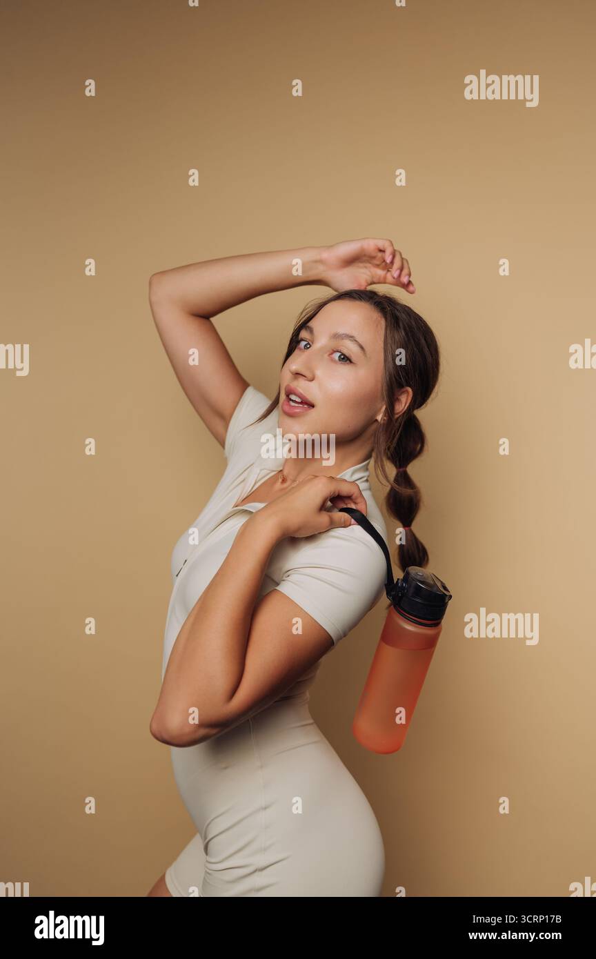 Eine junge Frau posiert selbstbewusst mit einer Hand auf der Hüfte und einer anderen mit einer Wasserflasche. Sie zeigt ein sportliches Outfit in neutralen Farben Stockfoto