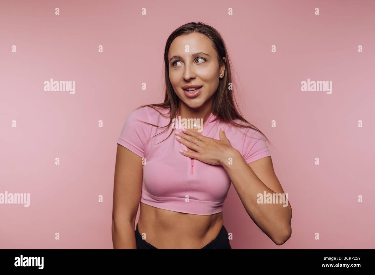Eine junge Frau steht selbstbewusst vor rosa Hintergrund, lächelt und legt eine Hand auf ihre Brust. Sie strahlt Energie und Verspieltheit in ihrem lässigen A aus Stockfoto