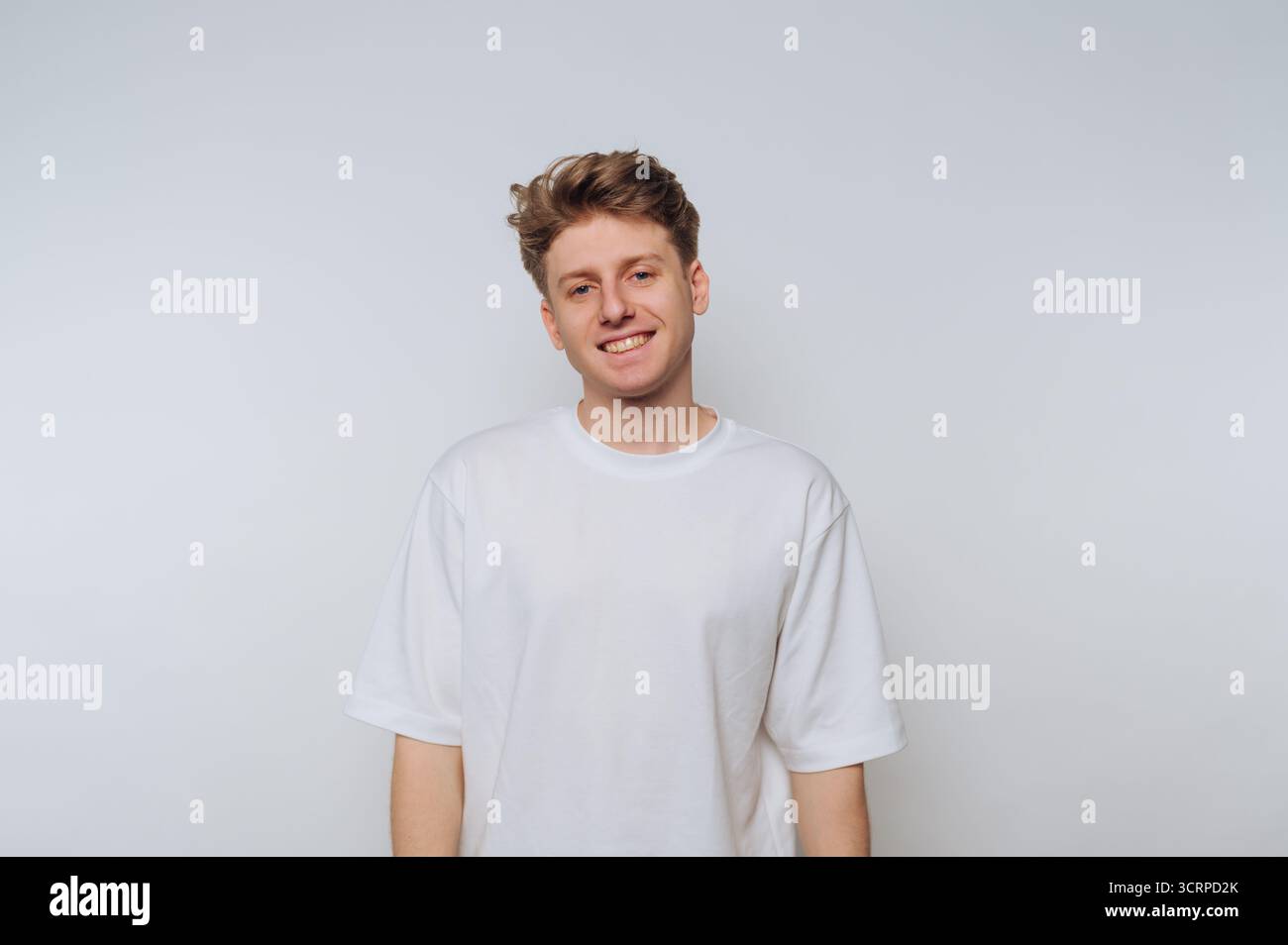 Ein junger Mann steht vor einem einfachen weißen Hintergrund und lächelt warm. Sein lässiges Outfit und sein freundlicher Ausdruck sorgen für eine einladende Atmosphäre, perfekt für Stockfoto