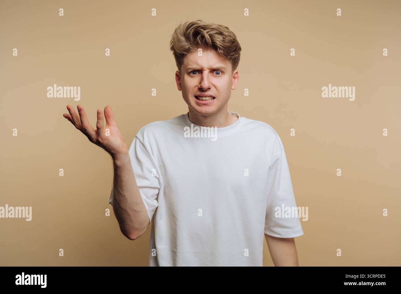 Der junge Mann steht vor beigefarbenem Hintergrund, sieht verwirrt aus mit erhobener Hand und zeigt eine Mischung aus Verwirrung und Frustration. Seine Freizeitkleidung Stockfoto