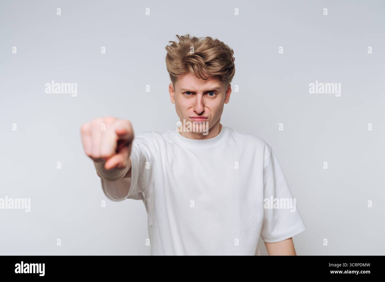 Ein junger Mann steht vor einem schlichten Hintergrund, sichtbar frustriert, als er mit einem starken Ausdruck direkt auf die Kamera zeigt und seine mes betont Stockfoto Ein junger Mann steht vor einem schlichten Hintergrund, sichtbar frustriert, als er mit einem starken Ausdruck direkt auf die Kamera zeigt und seine mes betont Stockfoto