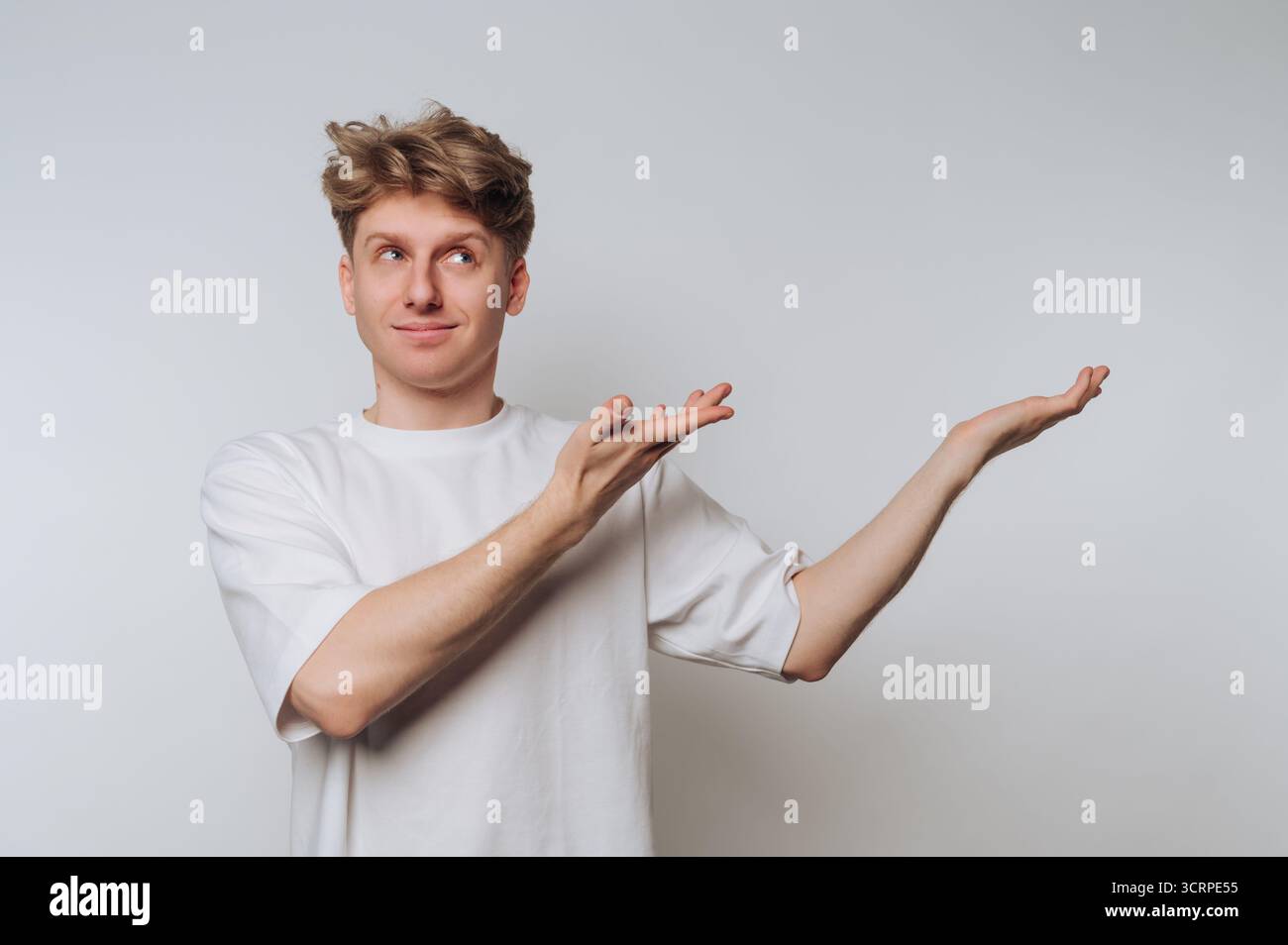 Ein junger Mann in einem lässigen weißen T-Shirt steht in einem schlichten Innenraum. Er hat einen verspielten Ausdruck, während er seine Arme nach außen streckt, als ob er einladend oder wäre Stockfoto