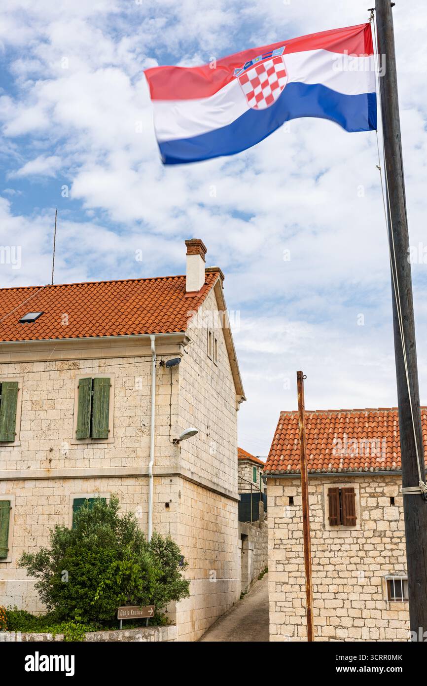 Eine kroatische Flagge fahnt über einem Steinhaus mit grünen Fensterläden und einem rot gekachelten Dach vor einem hellblauen Himmel. Eine heitere, ländliche Szene, die so toll ist Stockfoto