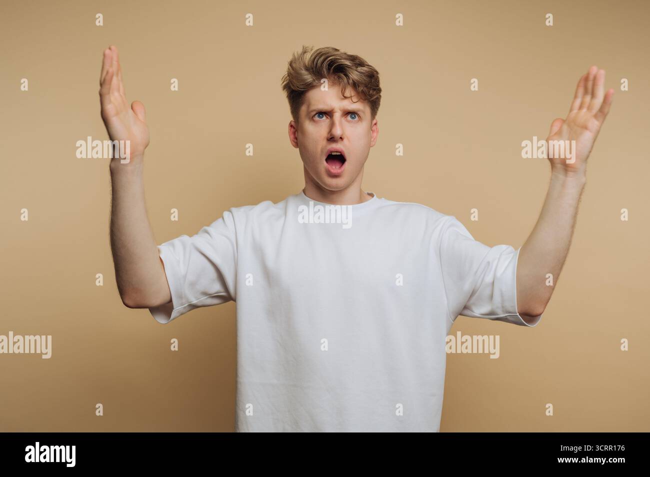 Ein junger Mann steht mit geöffnetem Mund und erhobenen Händen und zeigt einen starken Ausdruck von Überraschung oder Schock. Der Hintergrund ist ein warmer, neutraler Ton, enha Stockfoto