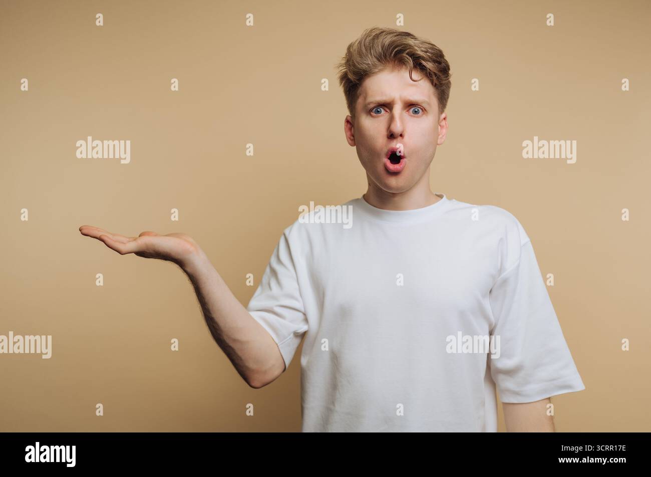 Ein junger Mann mit kurzen blonden Haaren zeigt einen überraschenden Ausdruck mit einer Geste der offenen Hand vor einem schlichten beigen Hintergrund. Seine Reaktion ca. Stockfoto
