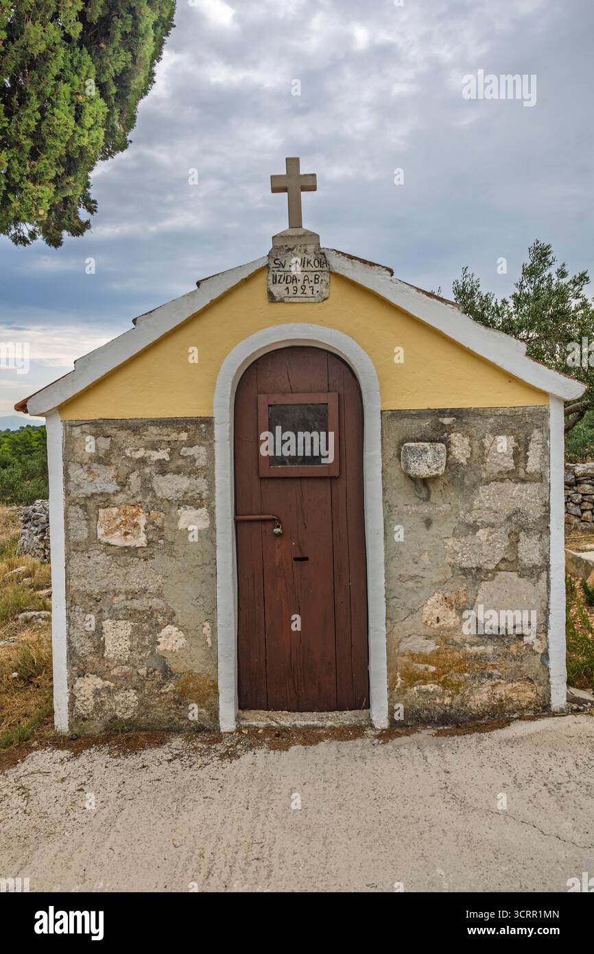 Eine kleine Steinkapelle mit gelbem Zierwerk und rot gekacheltem Dach befindet sich neben einer hohen Zypresse in einer sonnenüberfluteten ländlichen Landschaft. Die Szene ist leise Stockfoto