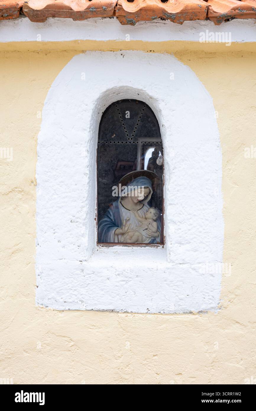 Ein kleines religiöses Bild von Maria mit Jesuskind in einer bogenförmigen Fensternische auf einer blassgelben Wand. Warmer, hingebungsvoller, rustikaler kirchlicher Charme mit V Stockfoto
