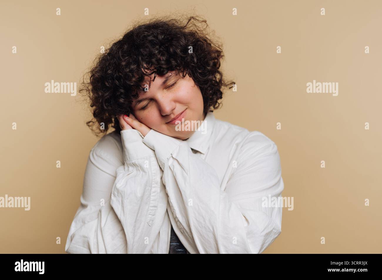 Eine Person mit lockigem Haar legt sich sanft auf die Hände und zeigt einen ruhigen und ruhigen Ausdruck. Der Hintergrund ist ein sanfter neutraler Ton, Stockfoto