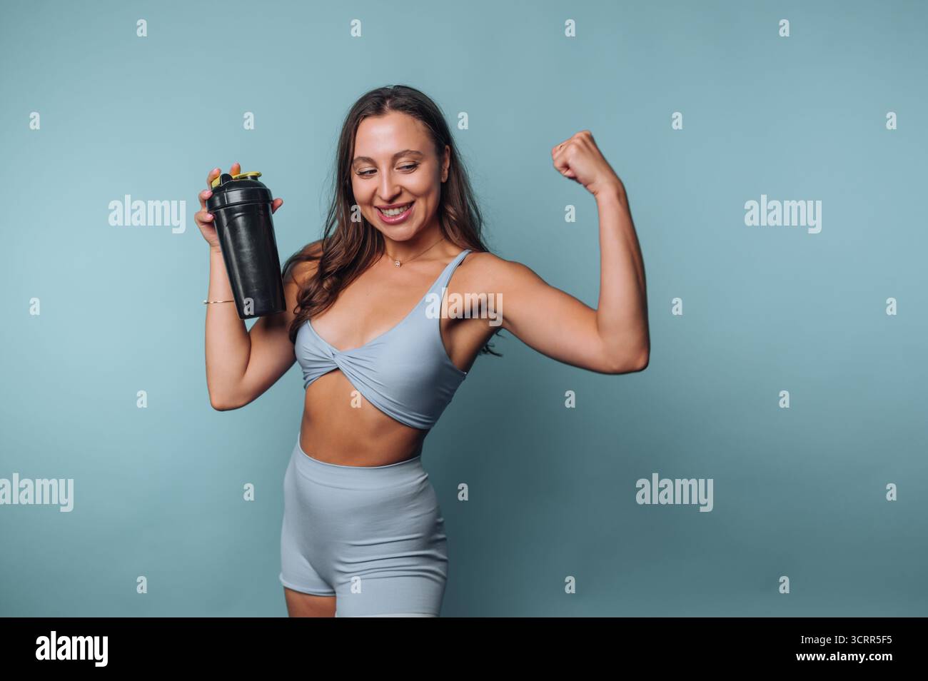 Eine selbstbewusste Frau in hellblauer Activewear beugt ihren Arm und zeigt Kraft, während sie einen Shaker hält. Stockfoto