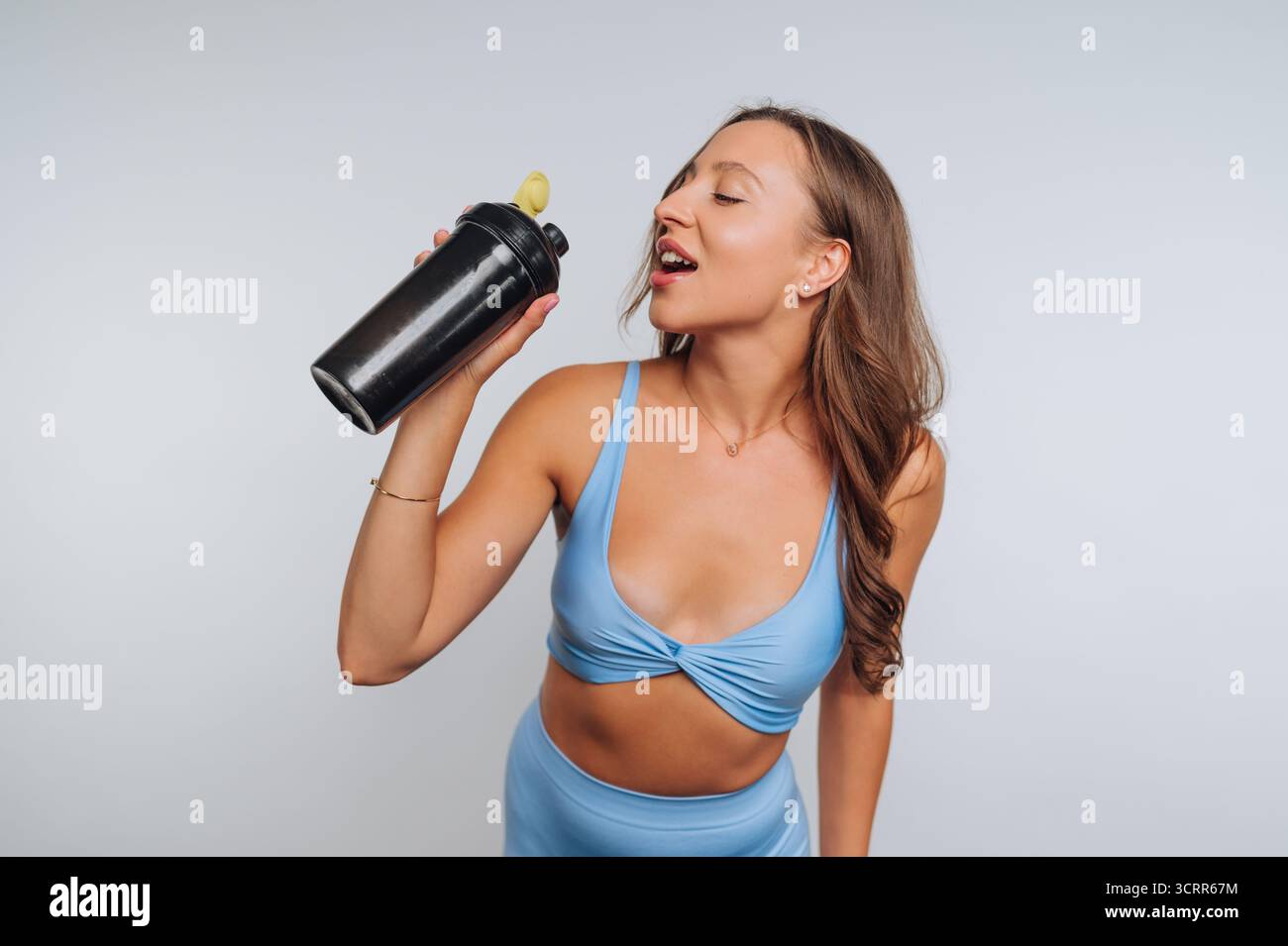 Eine Frau in einem hellblauen Workout-Outfit trinkt aus einer schwarzen Shaker-Flasche. Sie wirkt konzentriert und energiegeladen in ihrer Fitness-Routine. Der Hintergrund Stockfoto