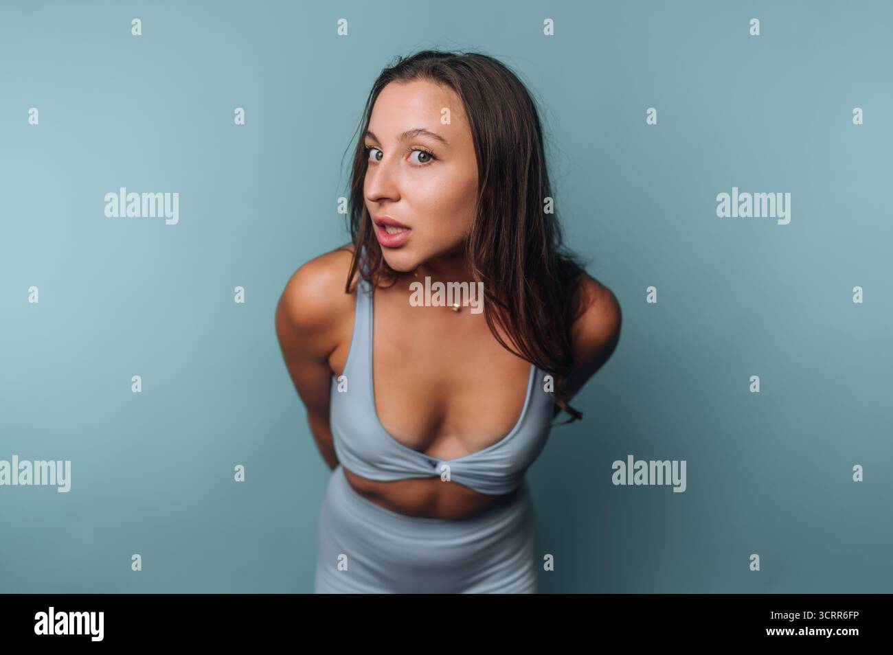 Eine junge Frau zeigt eine verspielte Pose, während sie ein hellblaues Sportoutfit trägt. Ihr Ausdruck vermittelt Selbstvertrauen vor einem passenden blauen Hintergrund. Stockfoto