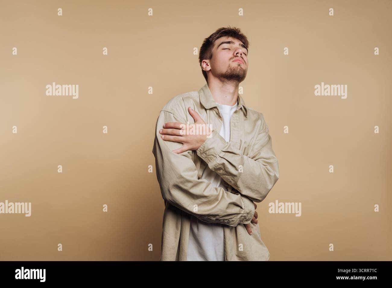 Ein junger Mann steht selbstbewusst vor einem neutralen Hintergrund, kreuzt seine Arme und zeigt eine entspannte, aber selbstbewusste Pose. Sein Outfit ist modern Stockfoto Ein junger Mann steht selbstbewusst vor einem neutralen Hintergrund, kreuzt seine Arme und zeigt eine entspannte, aber selbstbewusste Pose. Sein Outfit ist modern Stockfoto