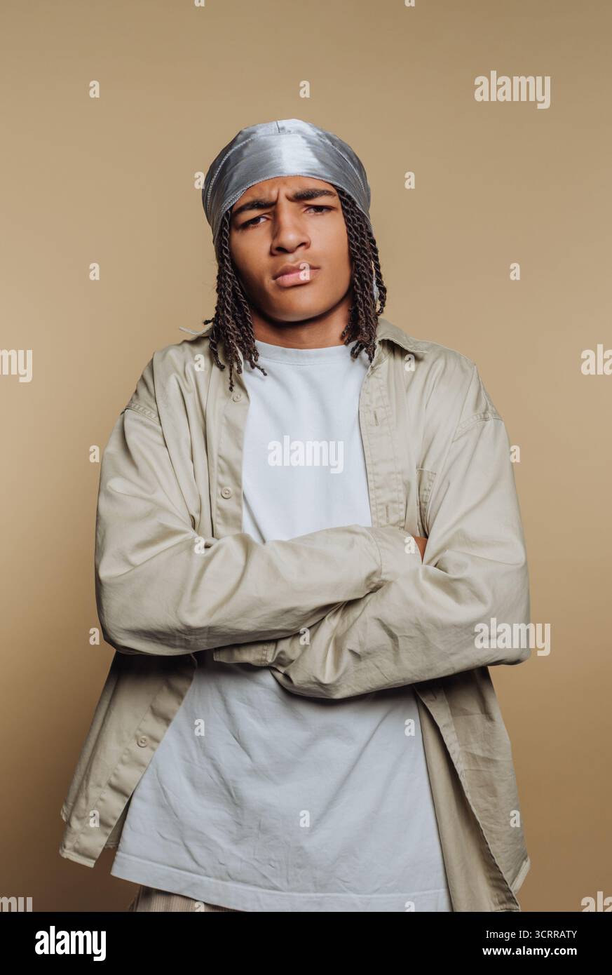 Junger Mann mit geflochtenen Haaren und silbernem durag, mit beigem Hemd und weißem T-Shirt, mit überkreuzten Armen vor beigem Hintergrund. Stockfoto