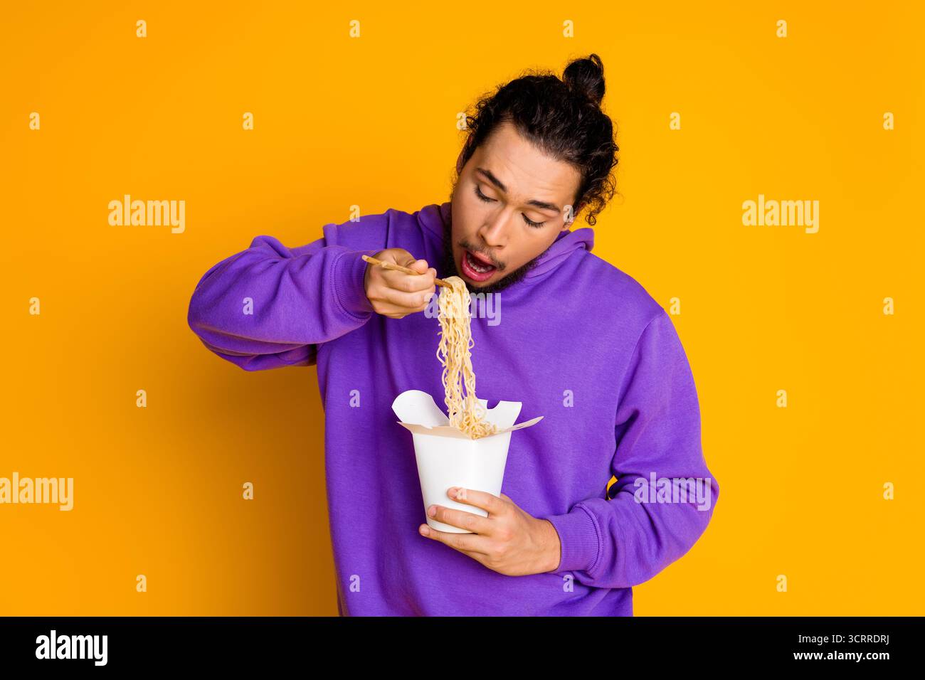 Porträt eines jungen Mannes in lässigem Hoodie, der Nudeln vor hellgelbem Hintergrund isst und Spaß und Lebensstil zum Ausdruck bringt Stockfoto