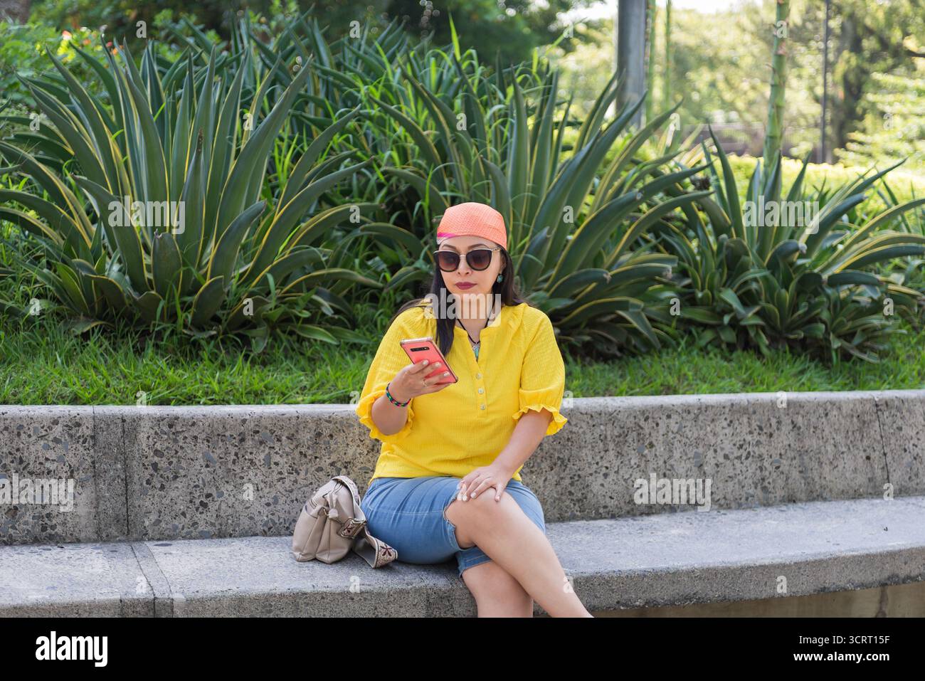 Eine Frau in gelbem Hemd und orangefarbenem Kopftuch sitzt auf einer Parkbank und benutzt ihr Telefon. Stockfoto
