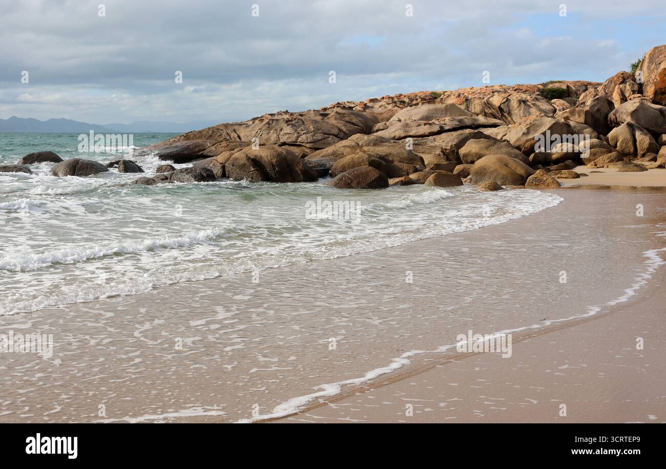 Sanfte Wellen, die sich an einem Sandstrand mit einem felsigen Felsvorsprung in der Rose Bay bei Bowen in Queensland, Australien, erheben Stockfoto