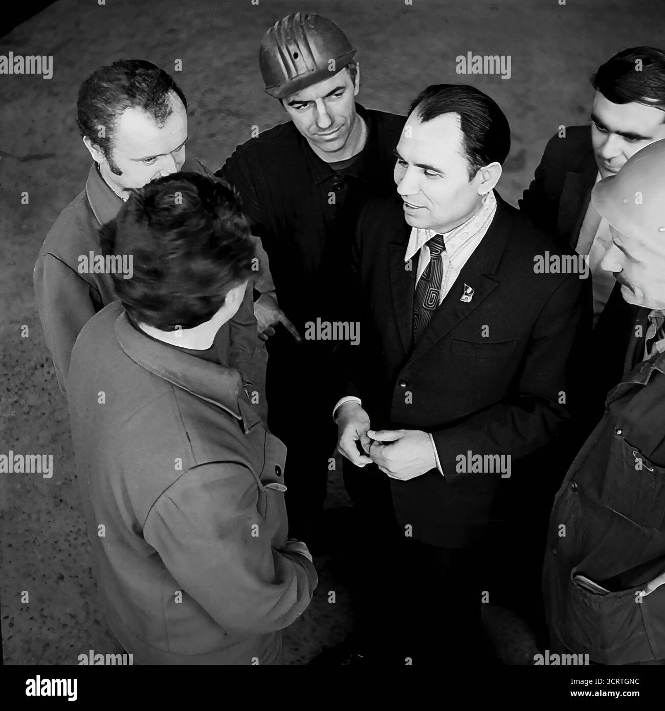 Ein Hochwinkelfoto aus dem Schwarzweiß-Archiv aus den 1970er Jahren zeigt einen sowjetischen Beamten oder Führer der Kommunistischen Partei, der sich mit Fabrikarbeitern in Sloviansk, der ukrainischen SSR, trifft. Der Beamte in einem formellen Anzug mit einem Staatsabzeichen am Revers steht in starkem Kontrast zu den Arbeitern in ihren Industriekleidung und Helmen. Dieses Bild ist eine klassische Darstellung des politischen Rituals der „Einheit der Partei und des Volkes“ und fängt einen Moment der Diskussion in der Fabrik während der Breschnew-Ära ein Stockfoto