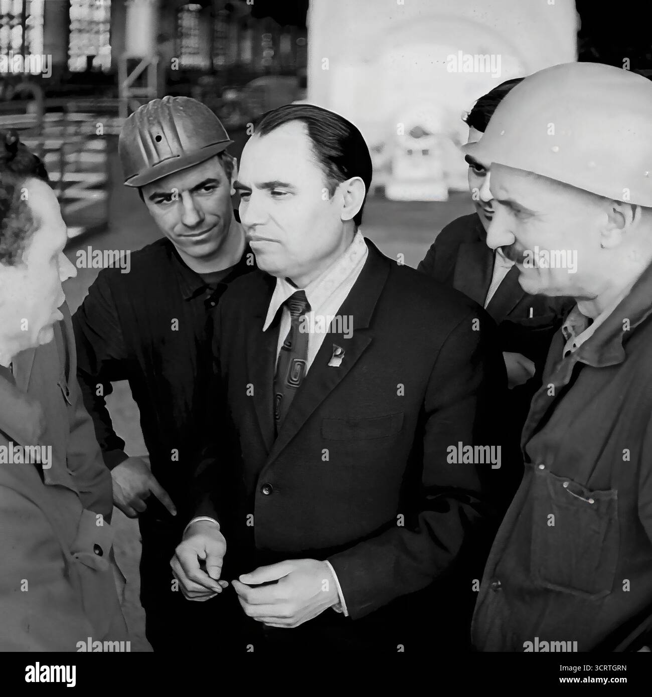 Ein Hochwinkelfoto aus dem Schwarzweiß-Archiv aus den 1970er Jahren zeigt einen sowjetischen Beamten oder Führer der Kommunistischen Partei, der sich mit Fabrikarbeitern in Sloviansk, der ukrainischen SSR, trifft. Der Beamte in einem formellen Anzug mit einem Staatsabzeichen am Revers steht in starkem Kontrast zu den Arbeitern in ihren Industriekleidung und Helmen. Dieses Bild ist eine klassische Darstellung des politischen Rituals der „Einheit der Partei und des Volkes“ und fängt einen Moment der Diskussion in der Fabrik während der Breschnew-Ära ein Stockfoto
