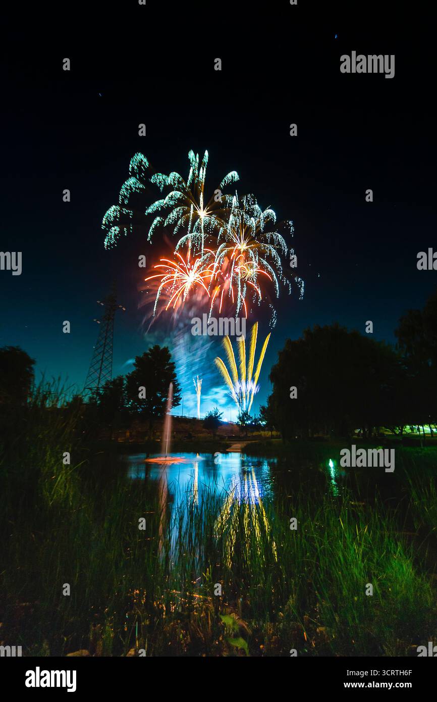 Buntes Feuerwerk erleuchtet den Nachthimmel und spiegelt sich auf der ruhigen Oberfläche eines Sees, der von Bäumen umgeben ist. Stockfoto