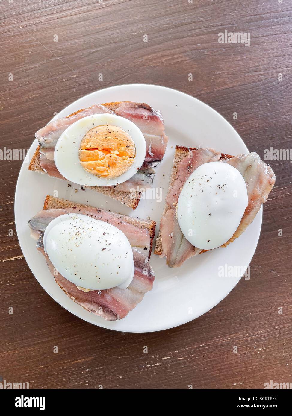 Sandwiches mit gesalzenen Sprotten und gekochten Ei werden auf Schwarzbrotscheiben serviert. Teller steht auf rustikalem Holztisch, bereit zum Essen Stockfoto