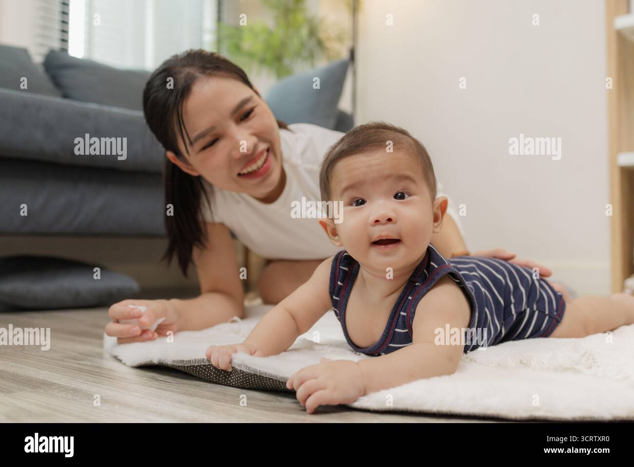 Ein Baby genießt die Bauchzeit auf einer weichen Matte, während die Mutter nah bleibt und es mit einem warmen Lächeln ermutigt. Dieser zärtliche Moment hebt frühe Entwicklungskräfte hervor Stockfoto