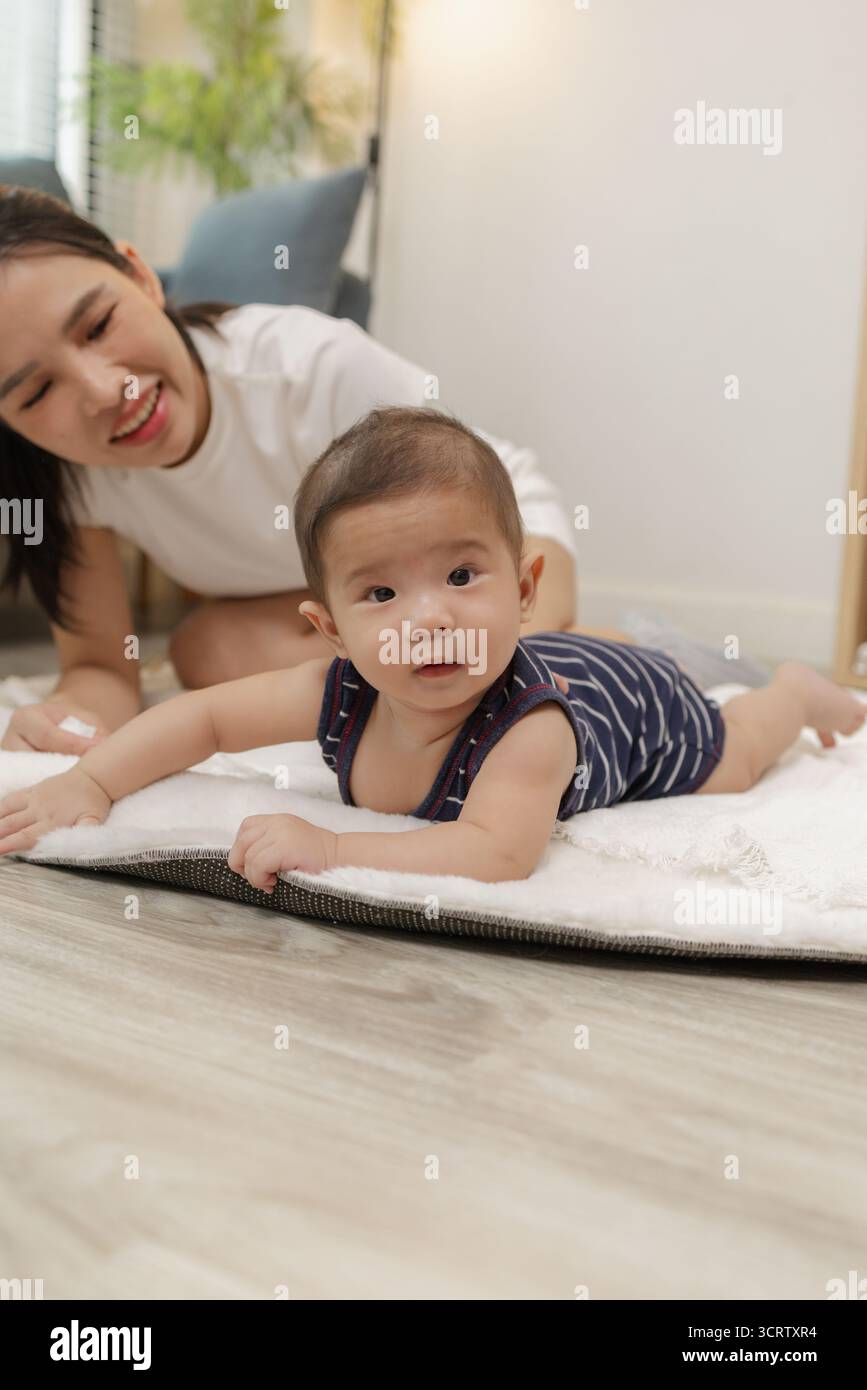 Ein Baby genießt die Bauchzeit auf einer weichen Matte, während die Mutter nah bleibt und es mit einem warmen Lächeln ermutigt. Dieser zärtliche Moment hebt frühe Entwicklungskräfte hervor Stockfoto