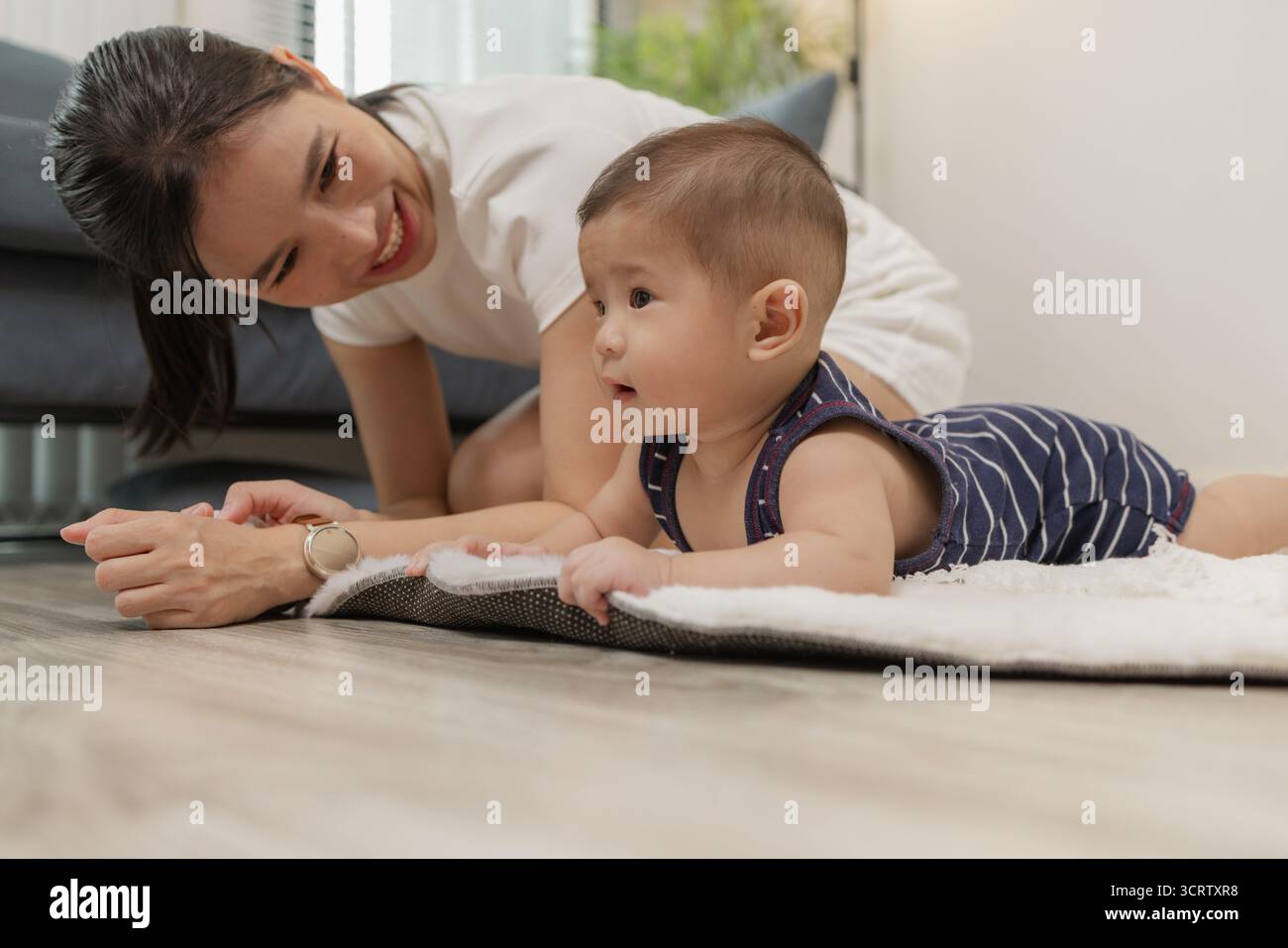 Ein Baby genießt die Bauchzeit auf einer weichen Matte, während die Mutter nah bleibt und es mit einem warmen Lächeln ermutigt. Dieser zärtliche Moment hebt frühe Entwicklungskräfte hervor Stockfoto