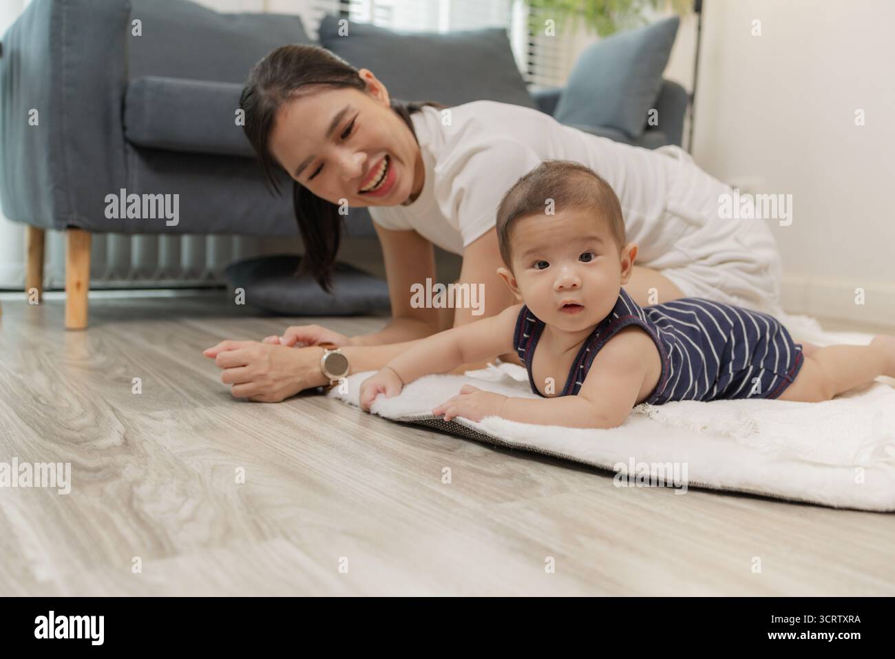 Ein Baby genießt die Bauchzeit auf einer weichen Matte, während die Mutter nah bleibt und es mit einem warmen Lächeln ermutigt. Dieser zärtliche Moment hebt frühe Entwicklungskräfte hervor Stockfoto