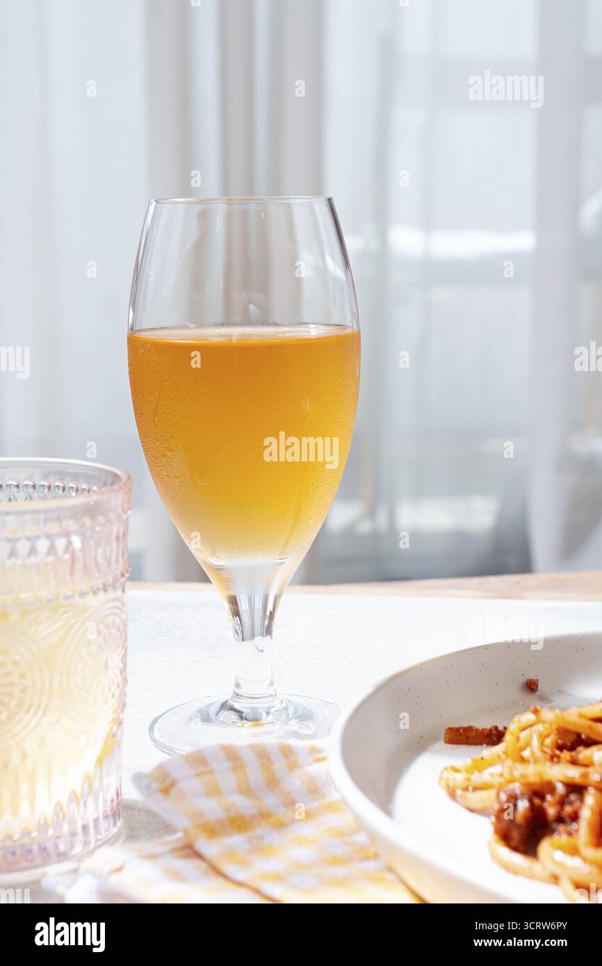Ein Glas mit einem hellen Bernsteingetränk steht neben einem Teller mit Nudeln. Helles Sonnenlicht strömt durch transparente Vorhänge und schafft einen warmen und einladenden geldautomaten Stockfoto