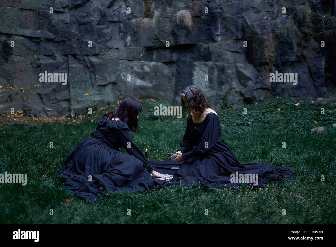 Zwei Frauen in langen schwarzen Kleidern sitzen an Halloween auf schwarzen Felsen, lesen Tarotkarten, führen ein Hexenritual durch, moderne Hexe Stockfoto