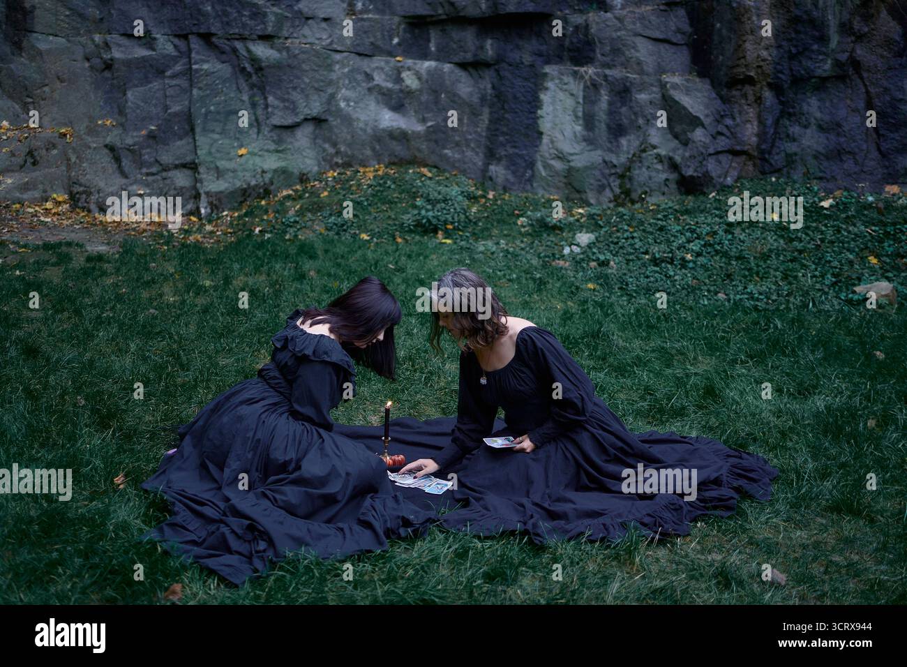 Zwei Frauen in langen schwarzen Kleidern sitzen an Halloween auf schwarzen Felsen, lesen Tarotkarten, führen ein Hexenritual durch, moderne Hexe Stockfoto